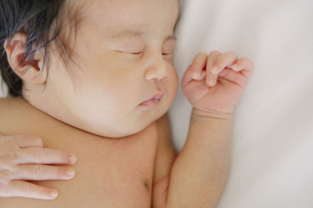 Peaceful newborn Asian baby sleeping on white blanket with tiny fist raised, resting on adult's shoulder.