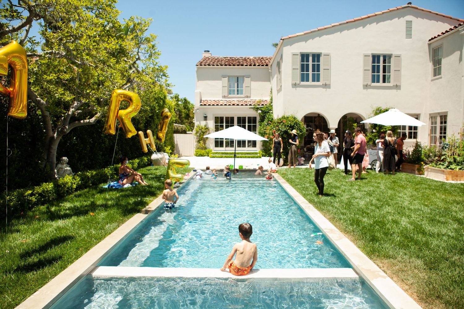 Children swimming in a backyard pool at a sunny outdoor party with gold balloon letters and guests.