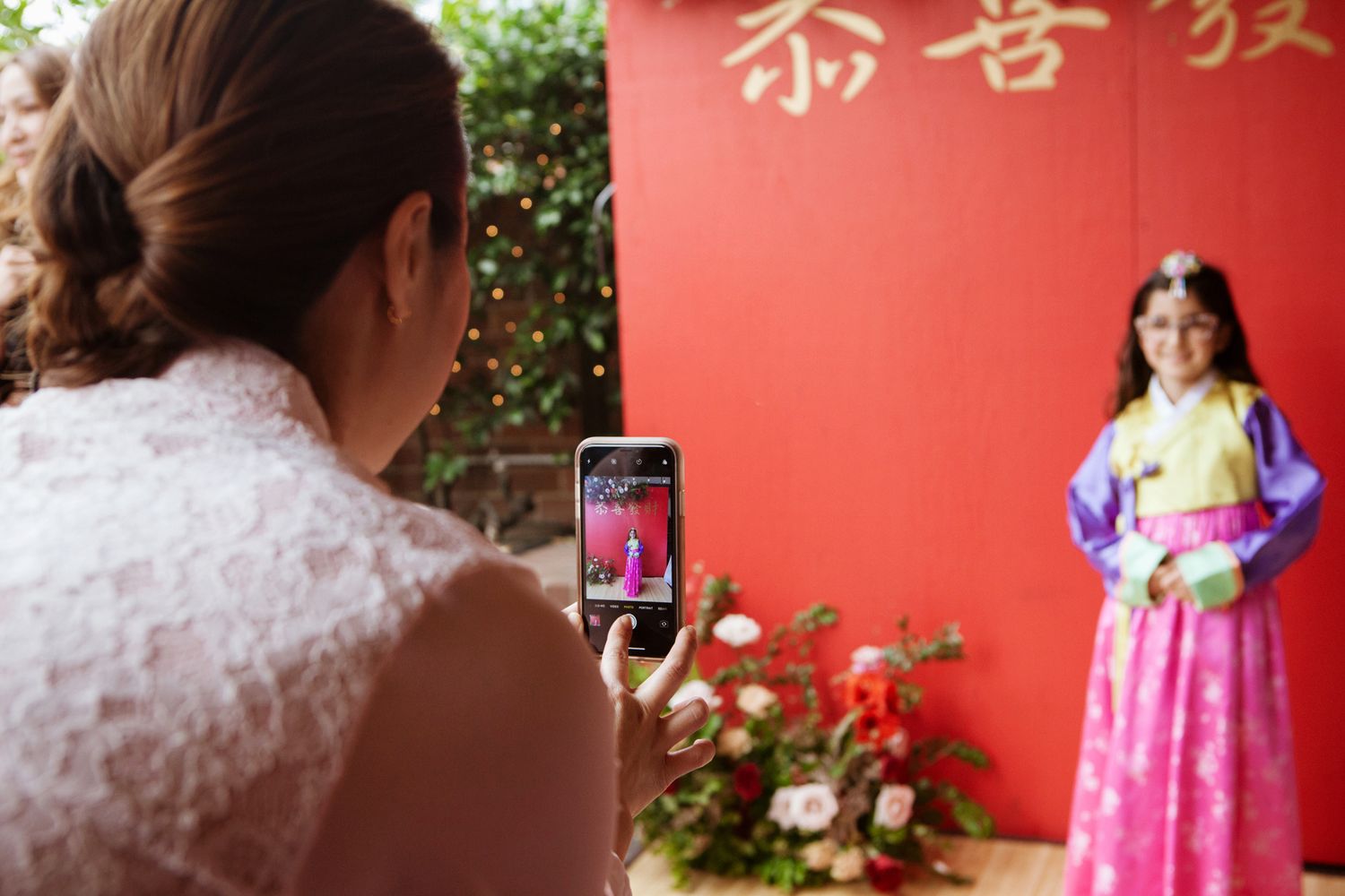 A woman photographs a girl posing in a pink Korean hanbok in front of a red Chinese New Year backdrop.