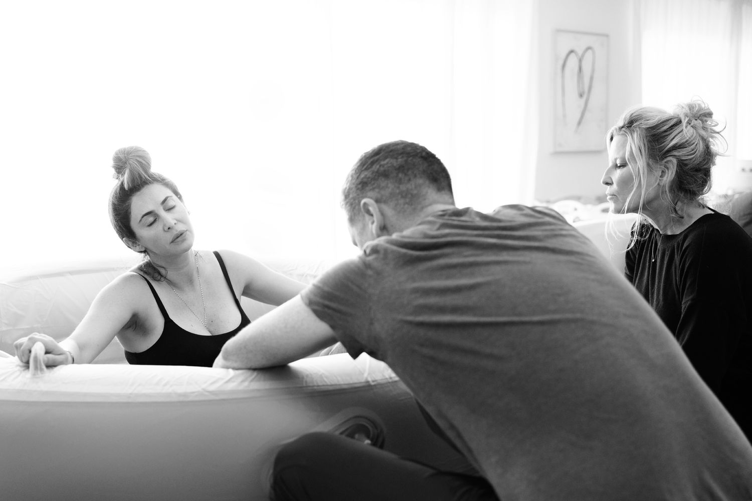 A midwife and partner support a laboring woman leaning on a birthing pool during an active home water birth.