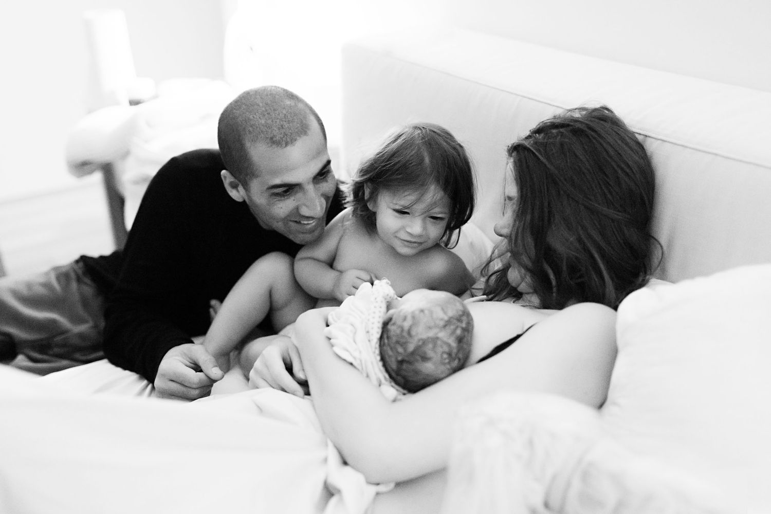Happy family with father, toddler, and mother admire newborn baby together on bed.