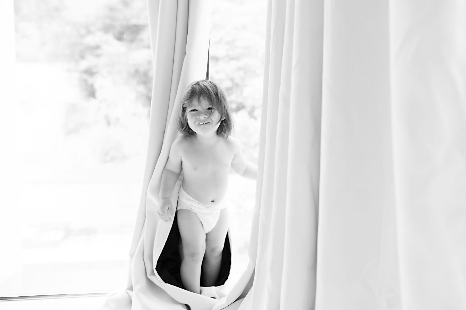 Black and white photo of a smiling toddler in a diaper peeking through white curtains near a bright window.