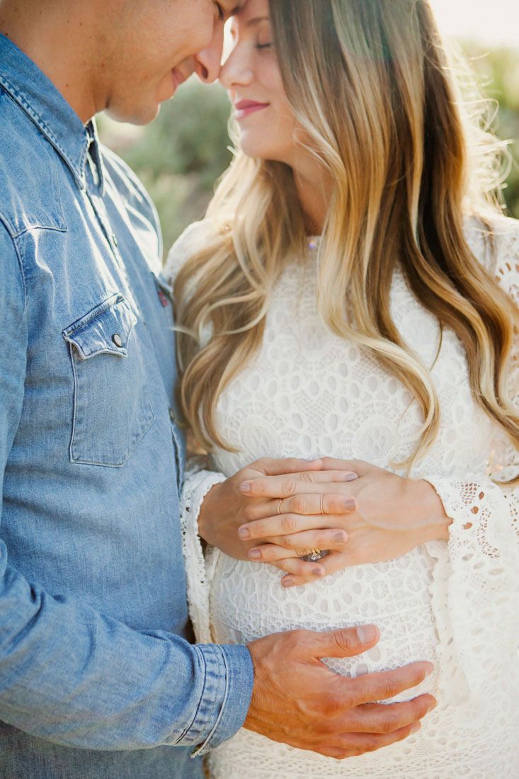 Couple tenderly touching pregnant belly outdoors, woman in white lace dress and man in denim shirt.