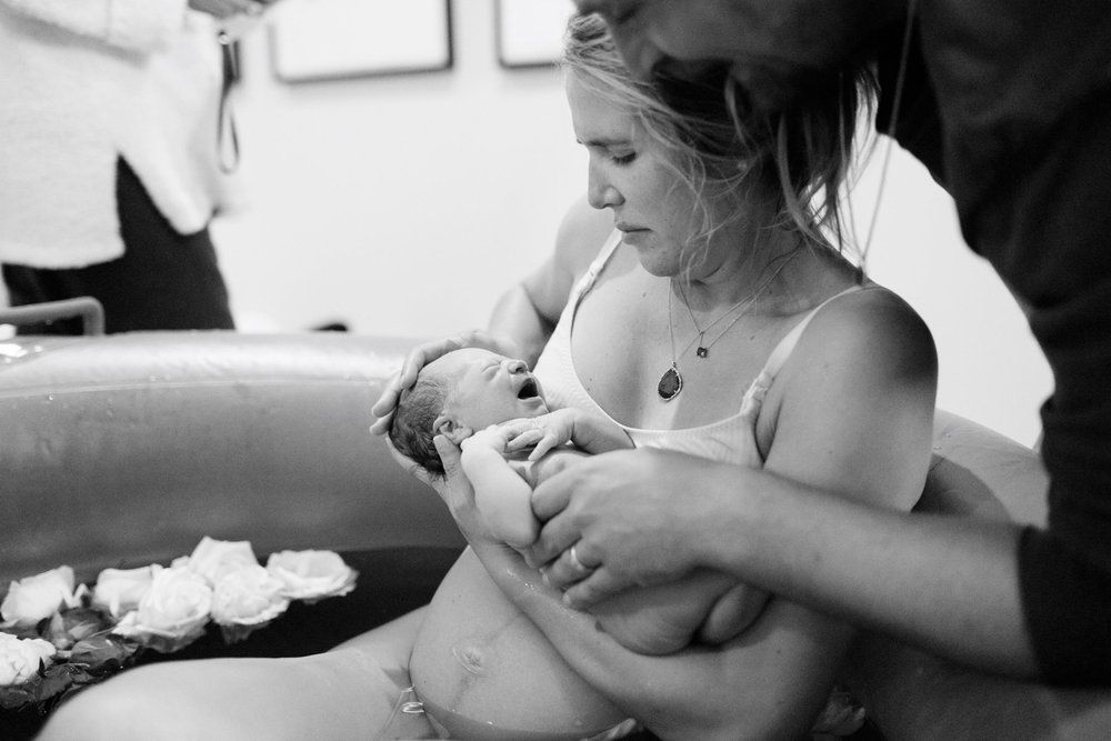 New mother cradling newborn in water birth tub surrounded by flowers, black and white.