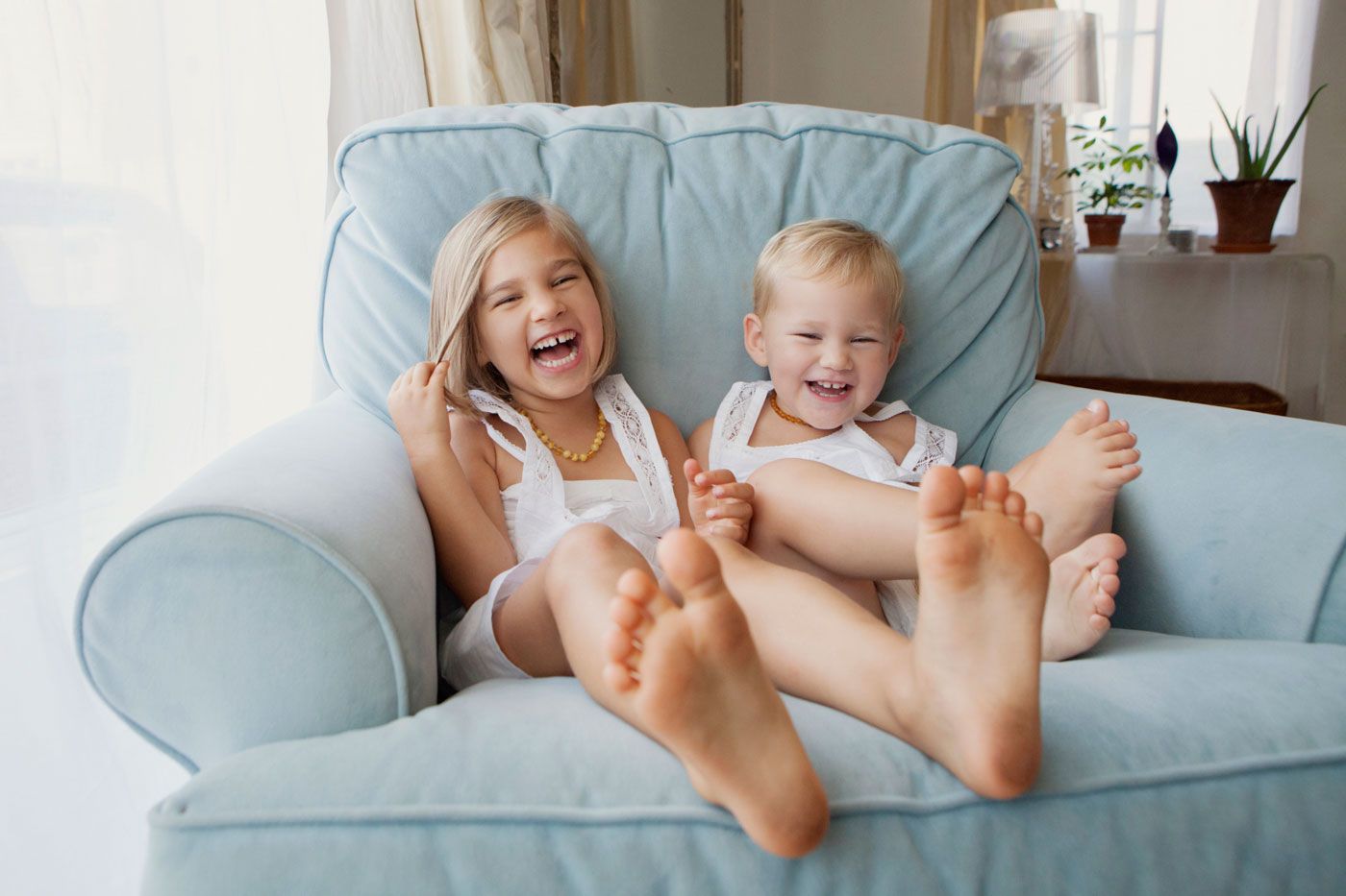 Two laughing young siblings sit barefoot together on a cozy blue armchair in a bright room.