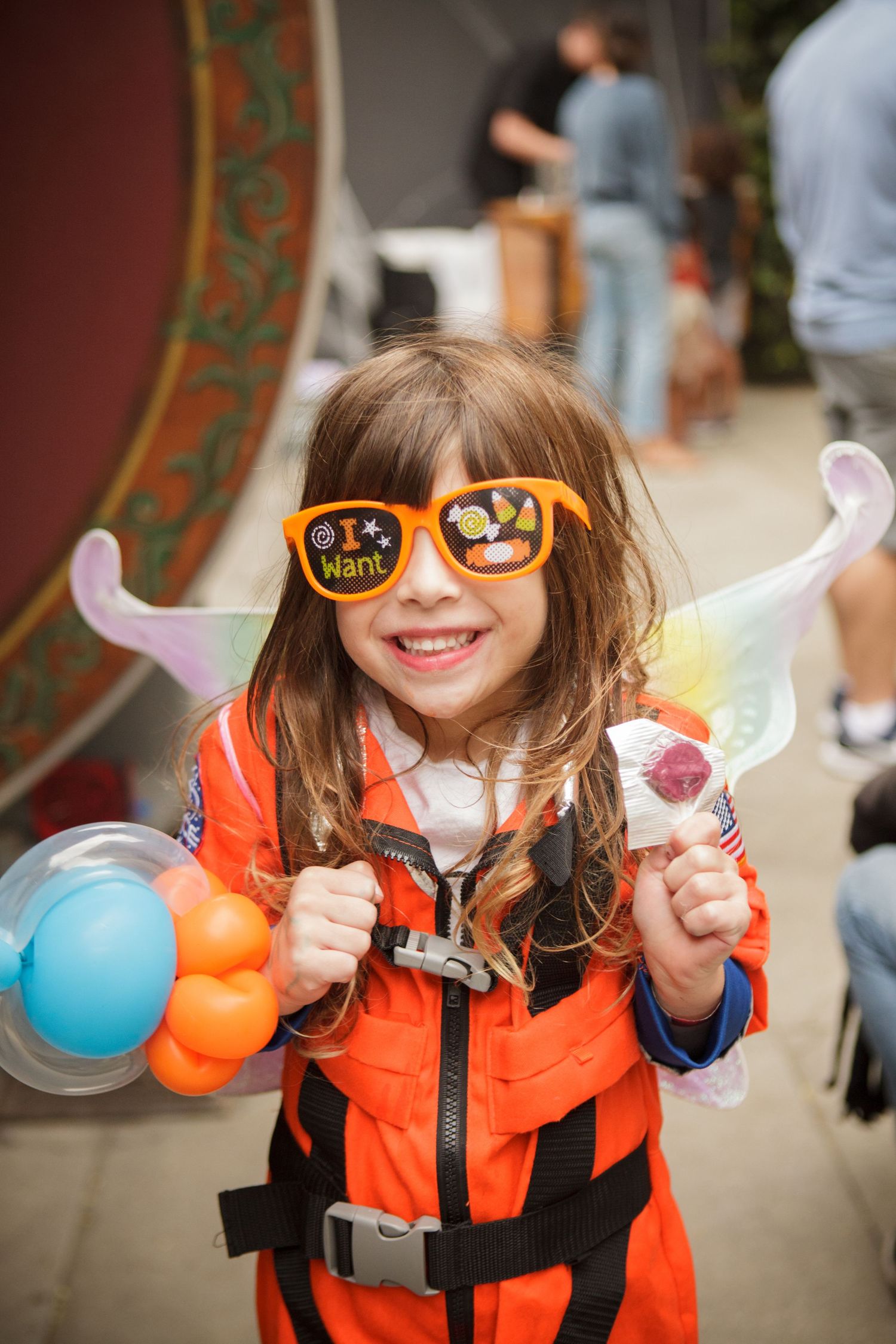 Happy young girl in orange jumpsuit wearing novelty sunglasses holding balloons and candy at a festive outdoor party.
