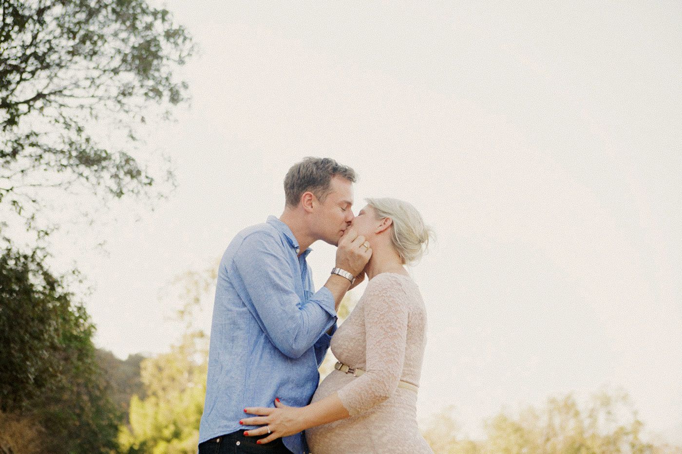 A couple shares a romantic kiss outdoors surrounded by lush green trees in warm natural light.