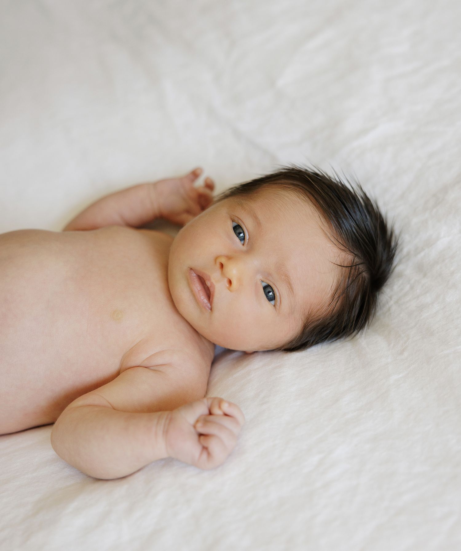 Newborn baby with dark hair lying on a white fluffy blanket, looking up with bright eyes.
