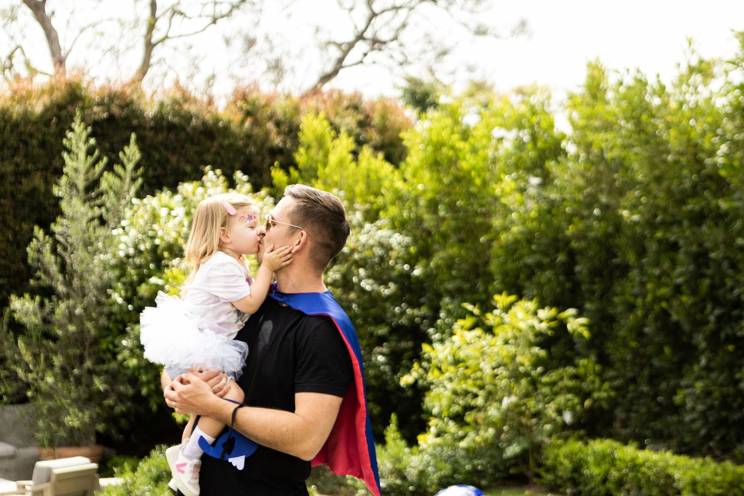 Young man in superhero cape tenderly kissing a toddler girl while holding her in a lush green garden.