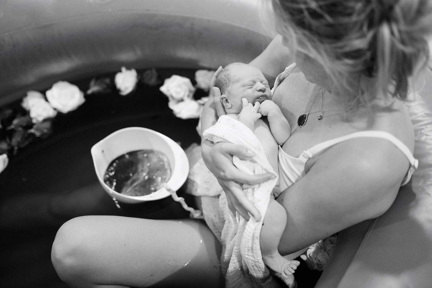Black and white image of a mother cradling her newborn in a water birth pool with floating white flowers.