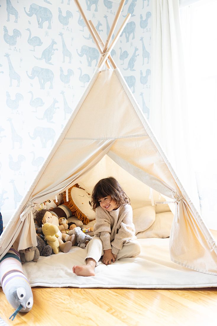 Happy young child sits inside a canvas teepee tent surrounded by stuffed animal toys indoors.