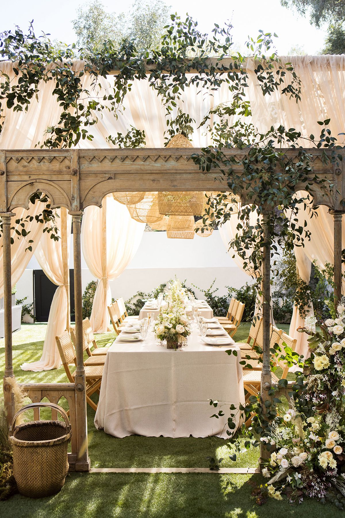 Beautiful outdoor wedding reception table set under a pergola draped with cream fabric and greenery.