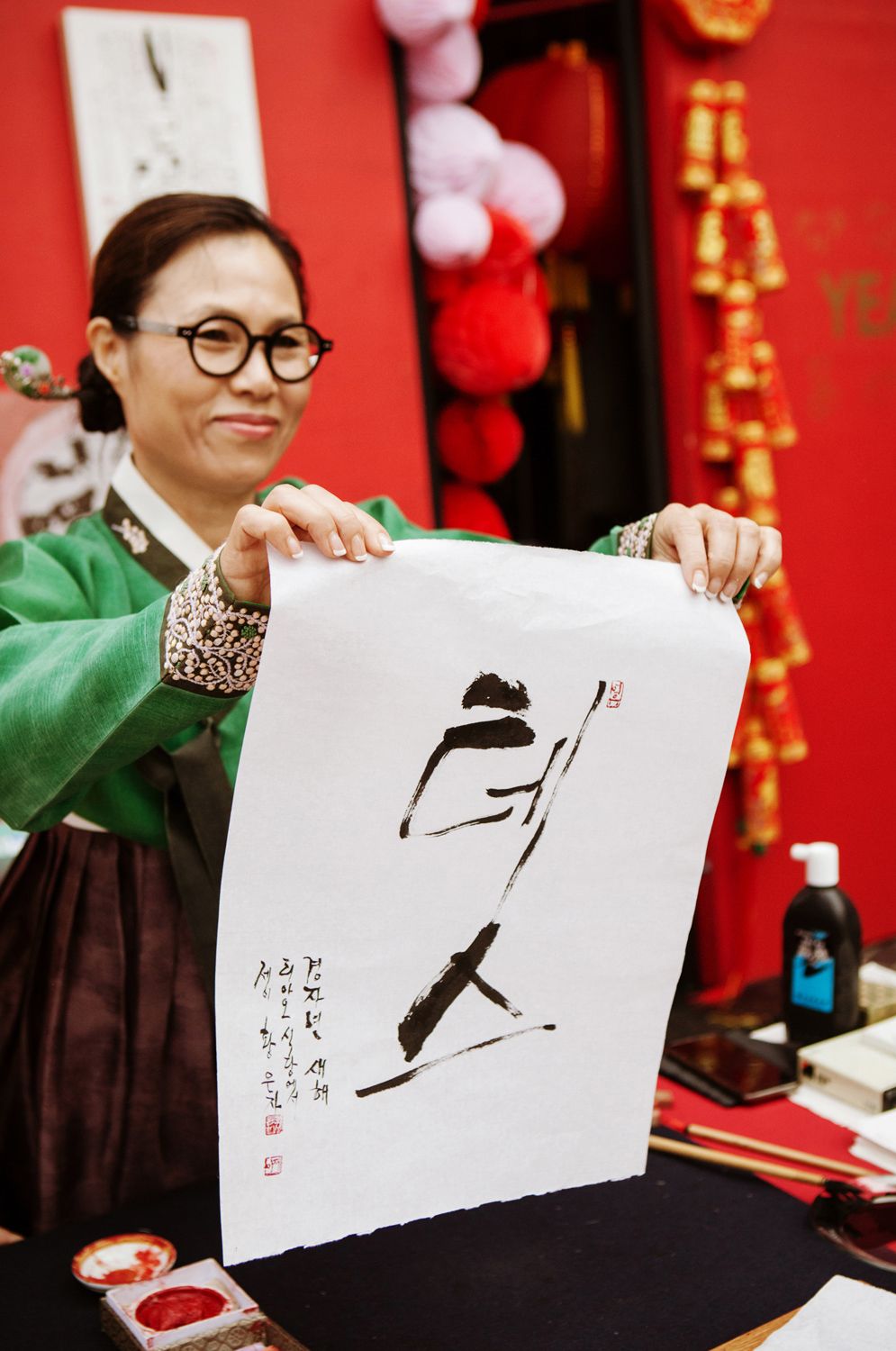 A woman in a green hanbok proudly displays a large piece of Korean calligraphy artwork at a cultural event.