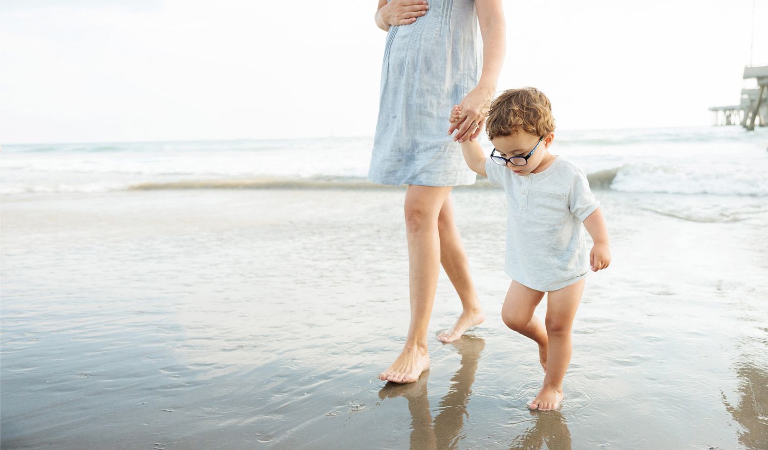 Young child in white outfit walking on wet sand at the beach with a pregnant mother nearby.