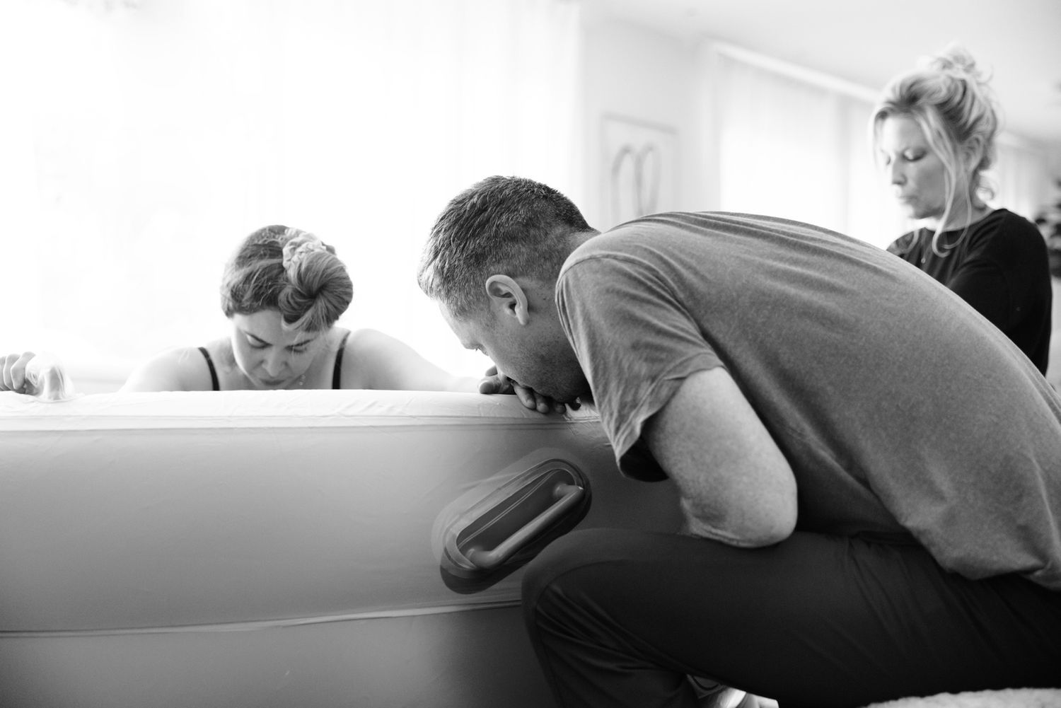 A partner and midwife kneel beside a birthing pool offering support during an active home water birth labor.