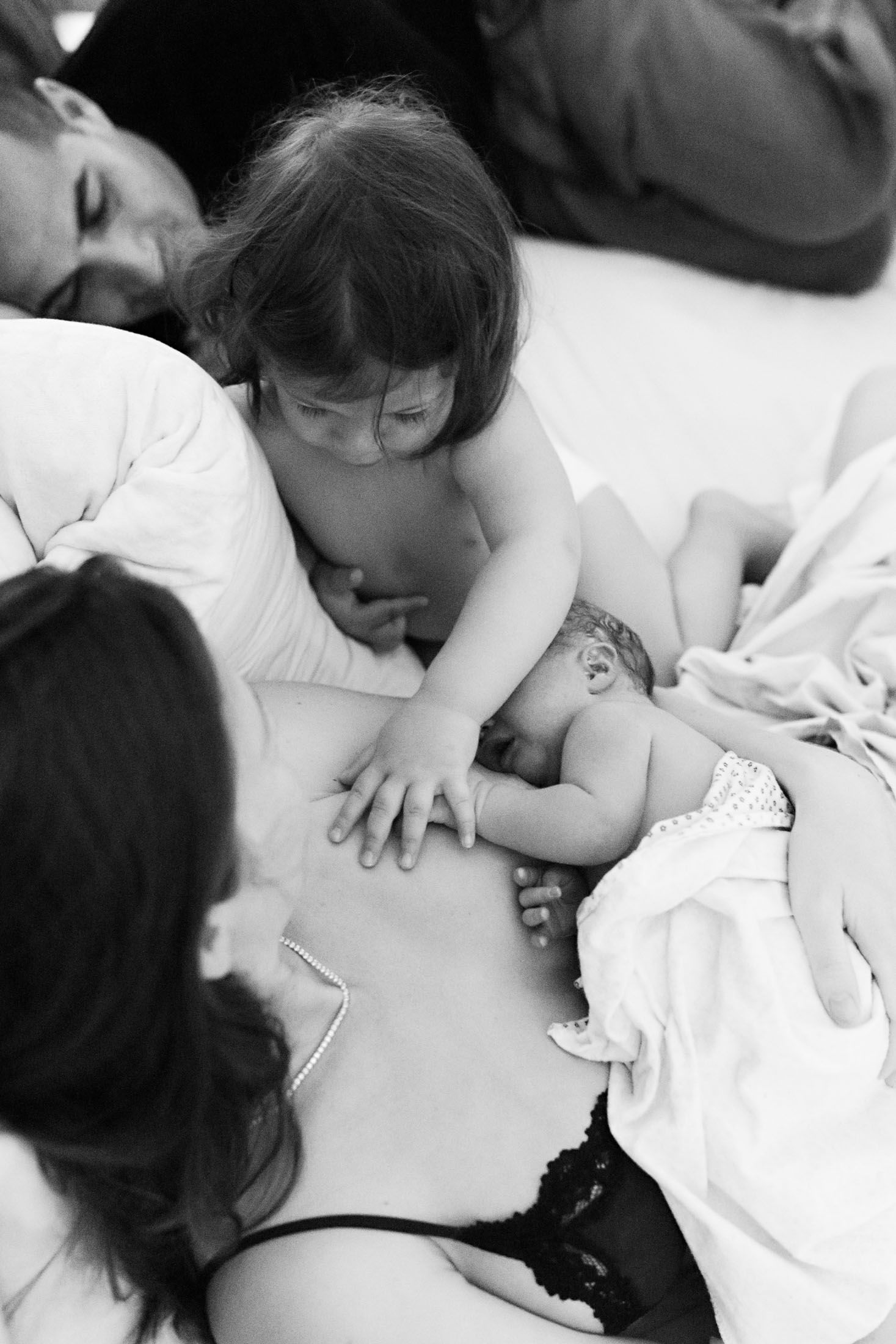 A toddler gently touches a newborn baby resting on a mothers chest in a tender black and white family photo.