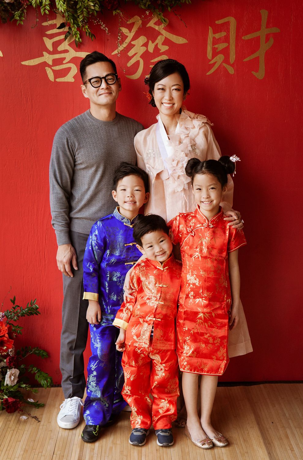 A family of five poses in traditional Chinese attire in front of a red Lunar New Year decorative backdrop.