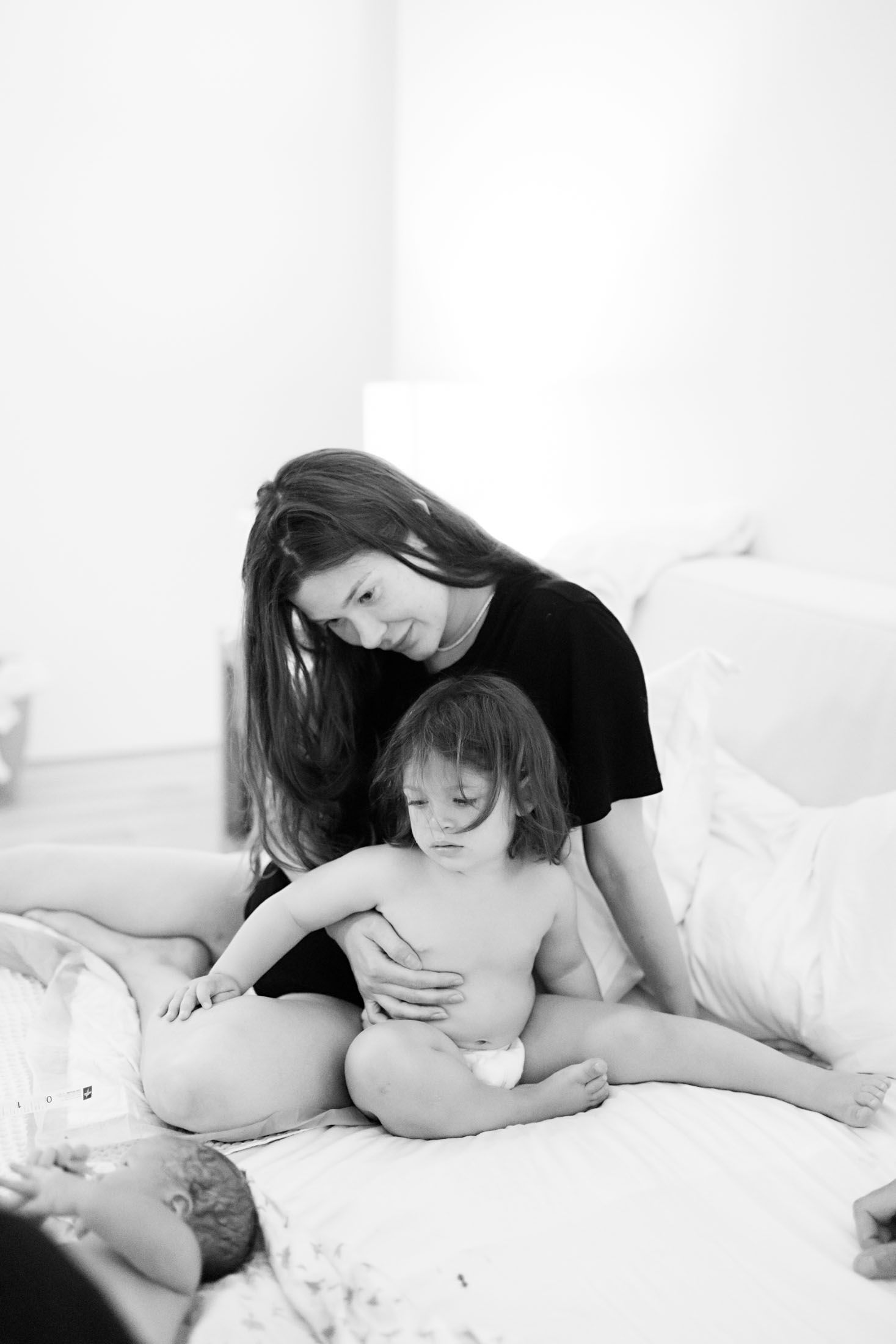 A mother in black sits behind her toddler on a white bed as they look at a newborn baby nearby.