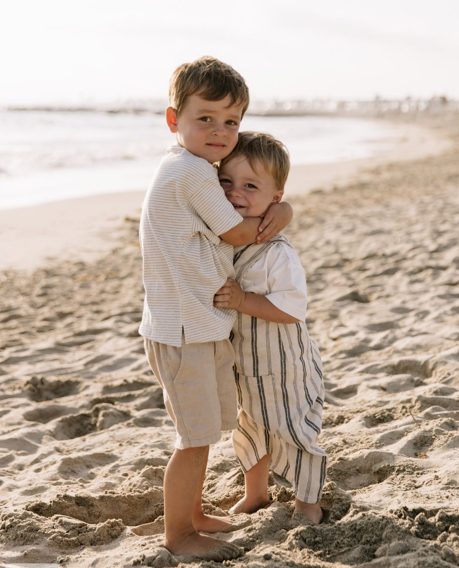 Two young boys hugging each other on a sandy beach, wearing neutral linen outfits, with ocean waves in the background.