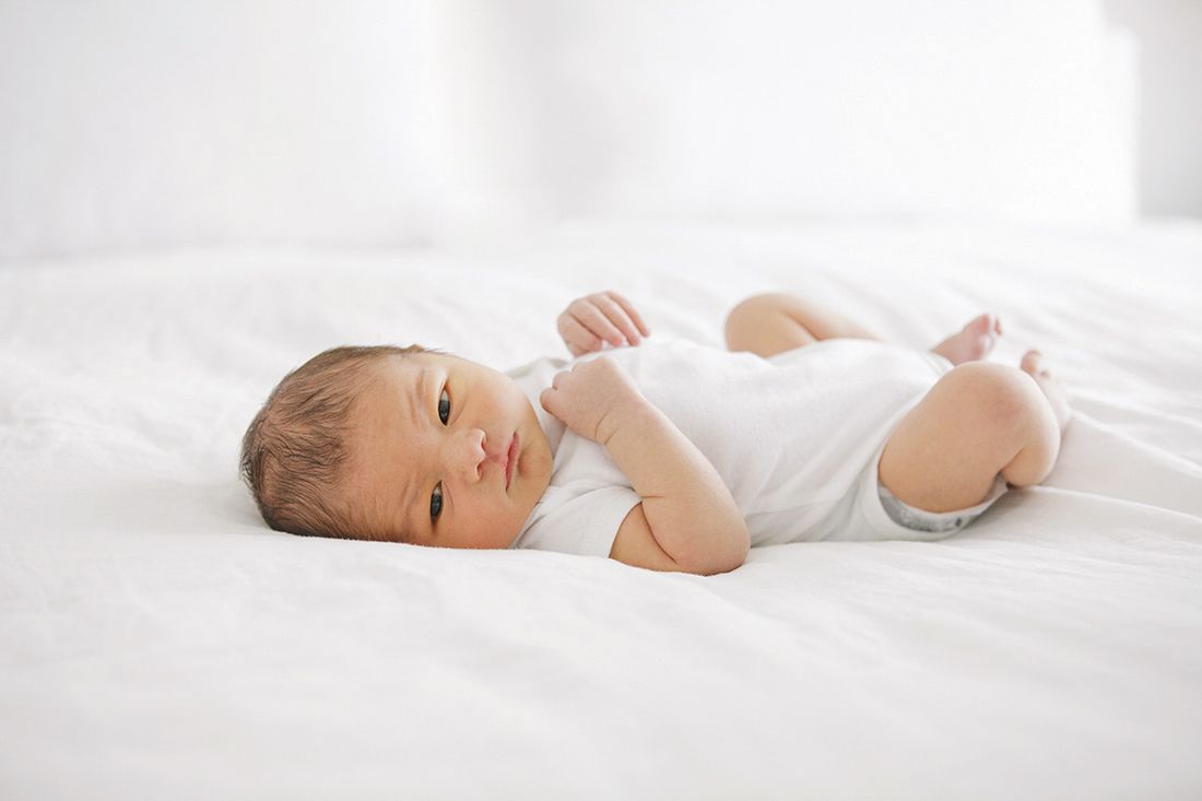 Newborn baby in white onesie lying on white bed, looking alert with dark hair and tiny hands curled up.