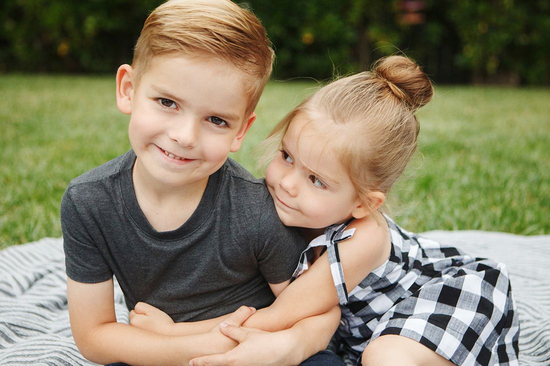 A smiling boy and girl sit together on a blanket in a lush green grassy yard.