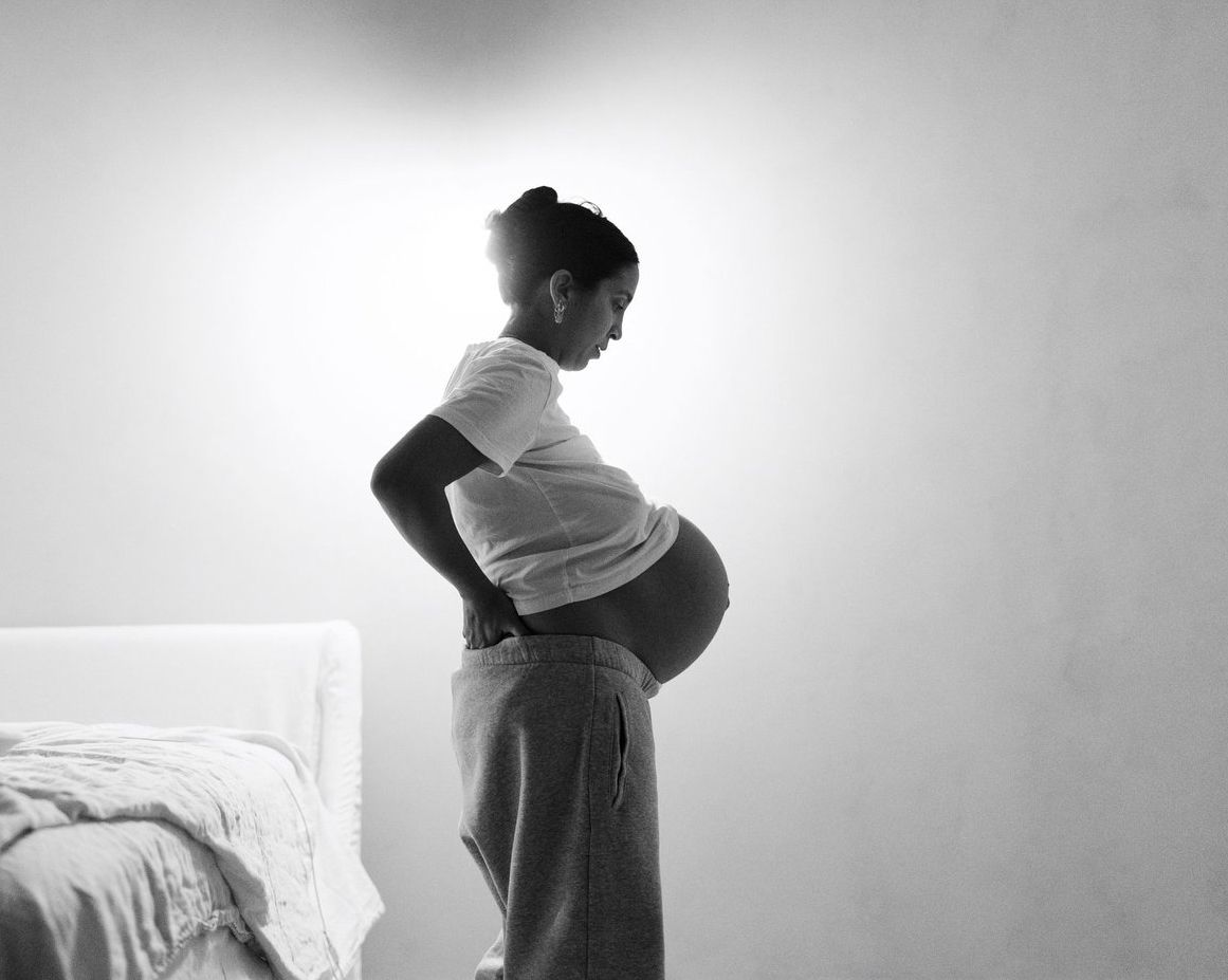 Black and white photo of a pregnant woman in profile, hands on hips, wearing a white top and gray sweatpants.