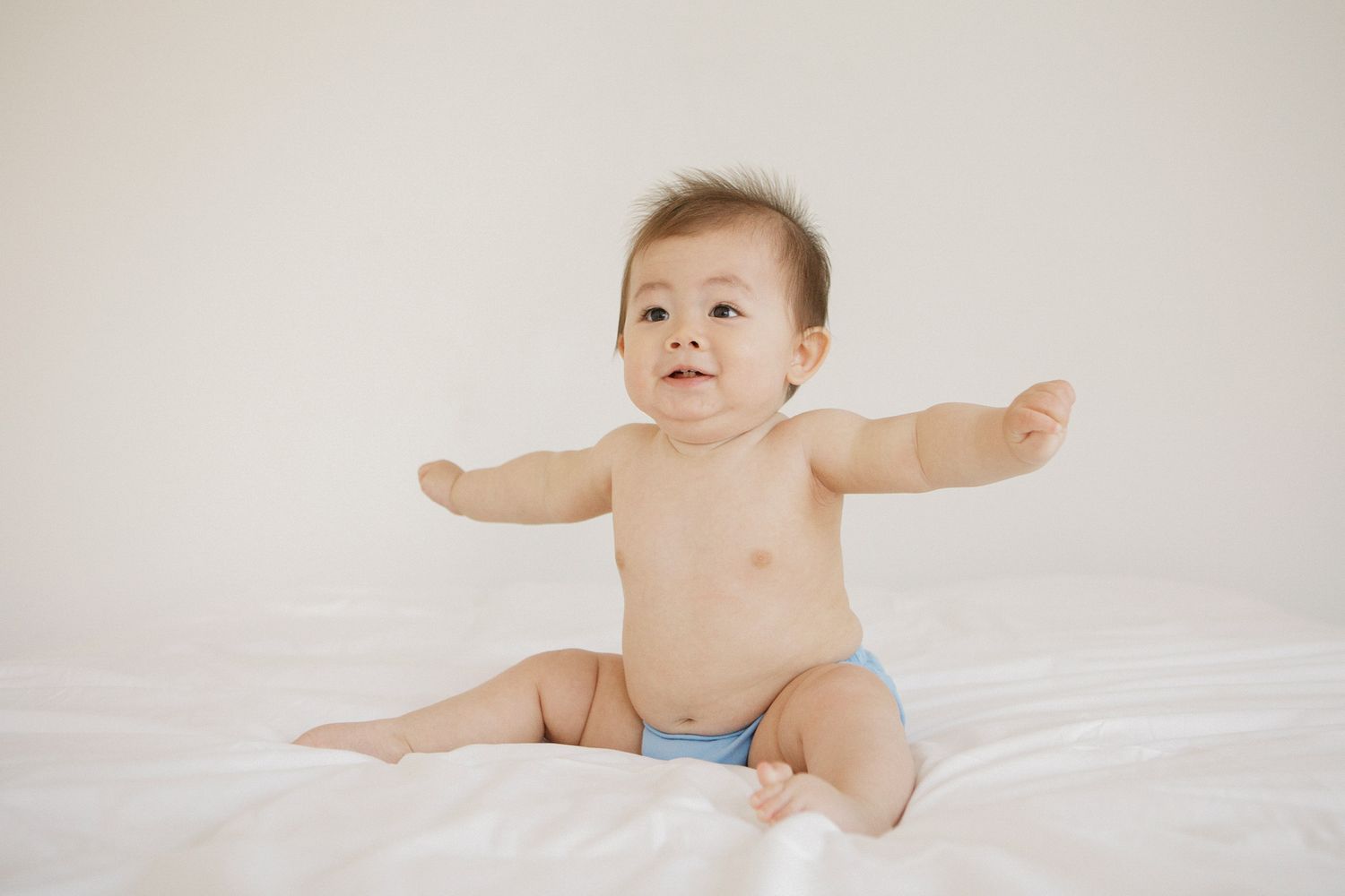 Happy baby sitting on white bed with arms outstretched wearing a blue diaper.