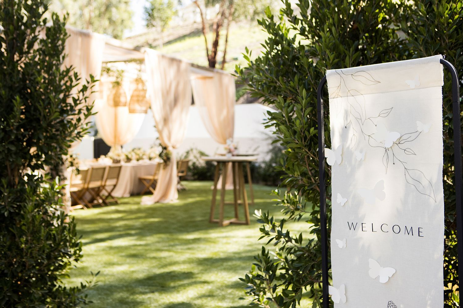 Elegant outdoor wedding welcome sign framed by lush greenery leading to a decorated reception area.