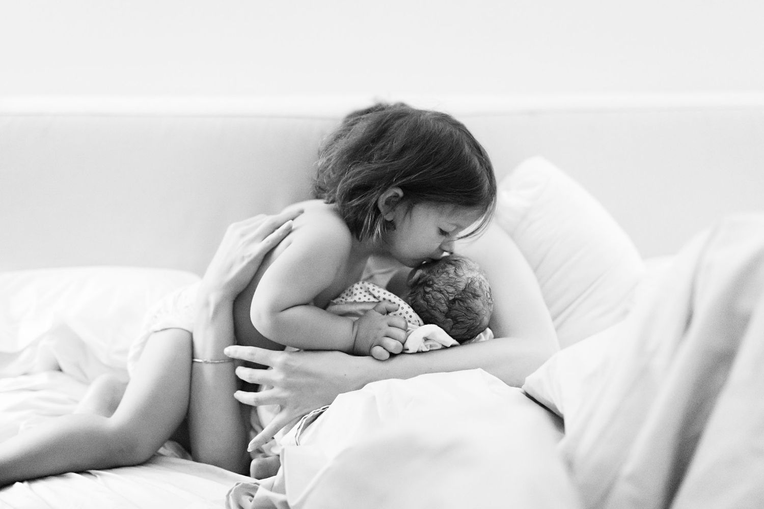 A young girl tenderly embraces a newborn baby while lying on a white bed in a black and white photo.