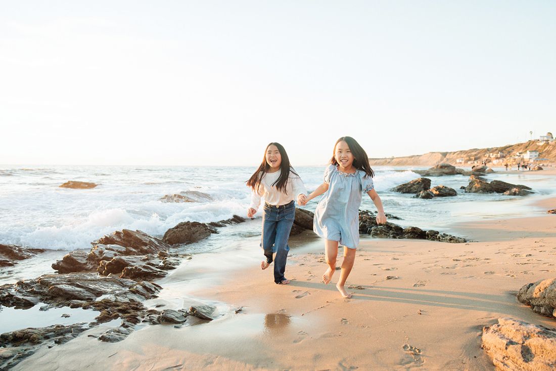Two young girls running and laughing together along a sunny rocky beach shoreline.