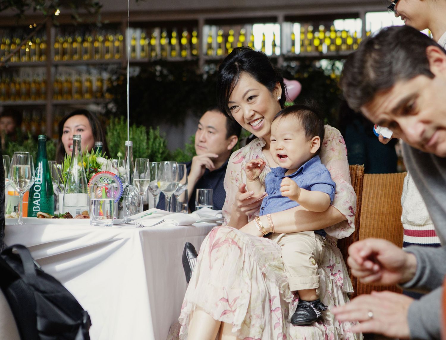 Smiling Asian woman holds crying baby at festive restaurant table during family celebration gathering.
