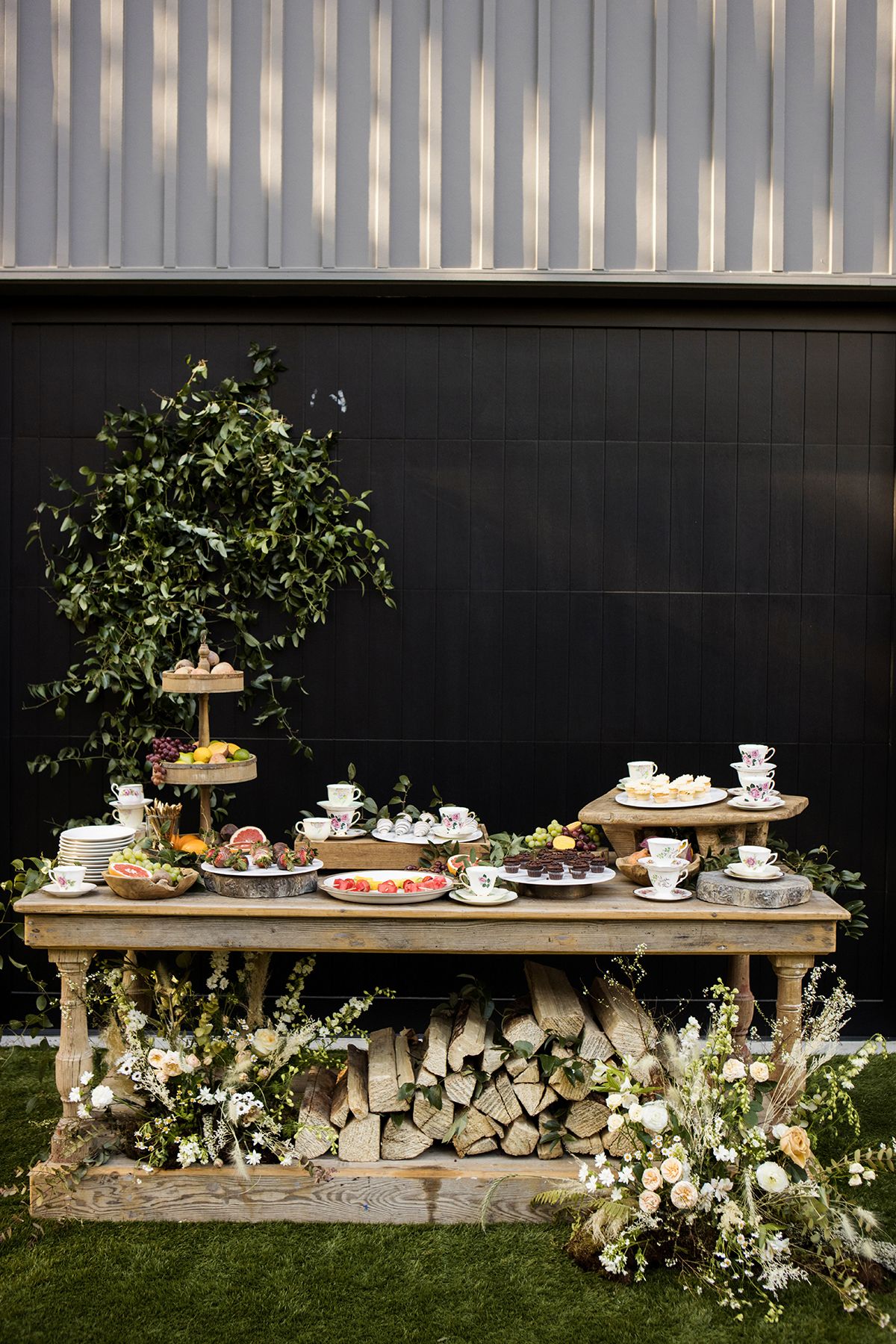 Rustic outdoor dessert table with tiered stands, teacups, and floral arrangements on a log-stacked base.