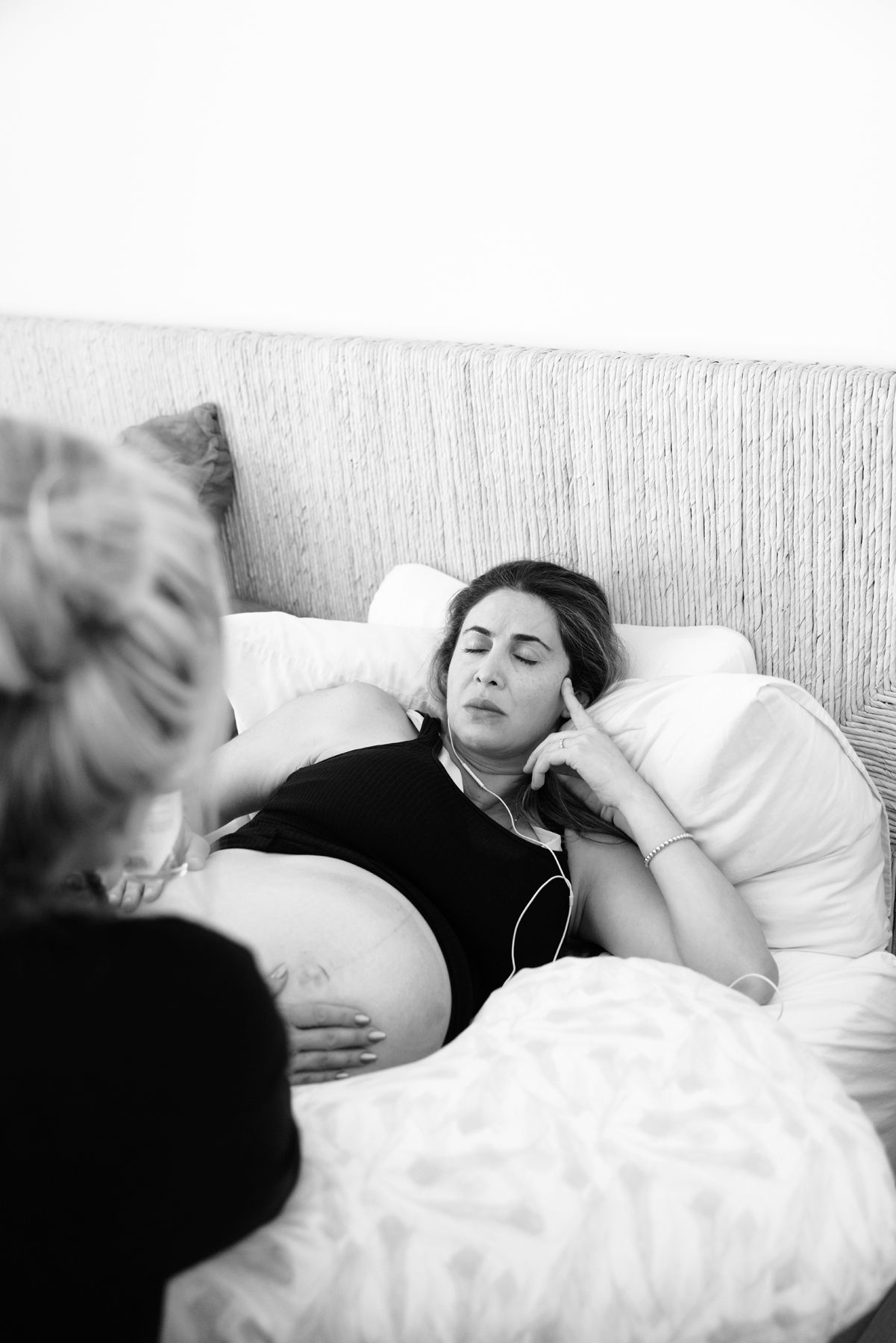 Pregnant woman resting on bed with earphones, receiving gentle belly support from doula.