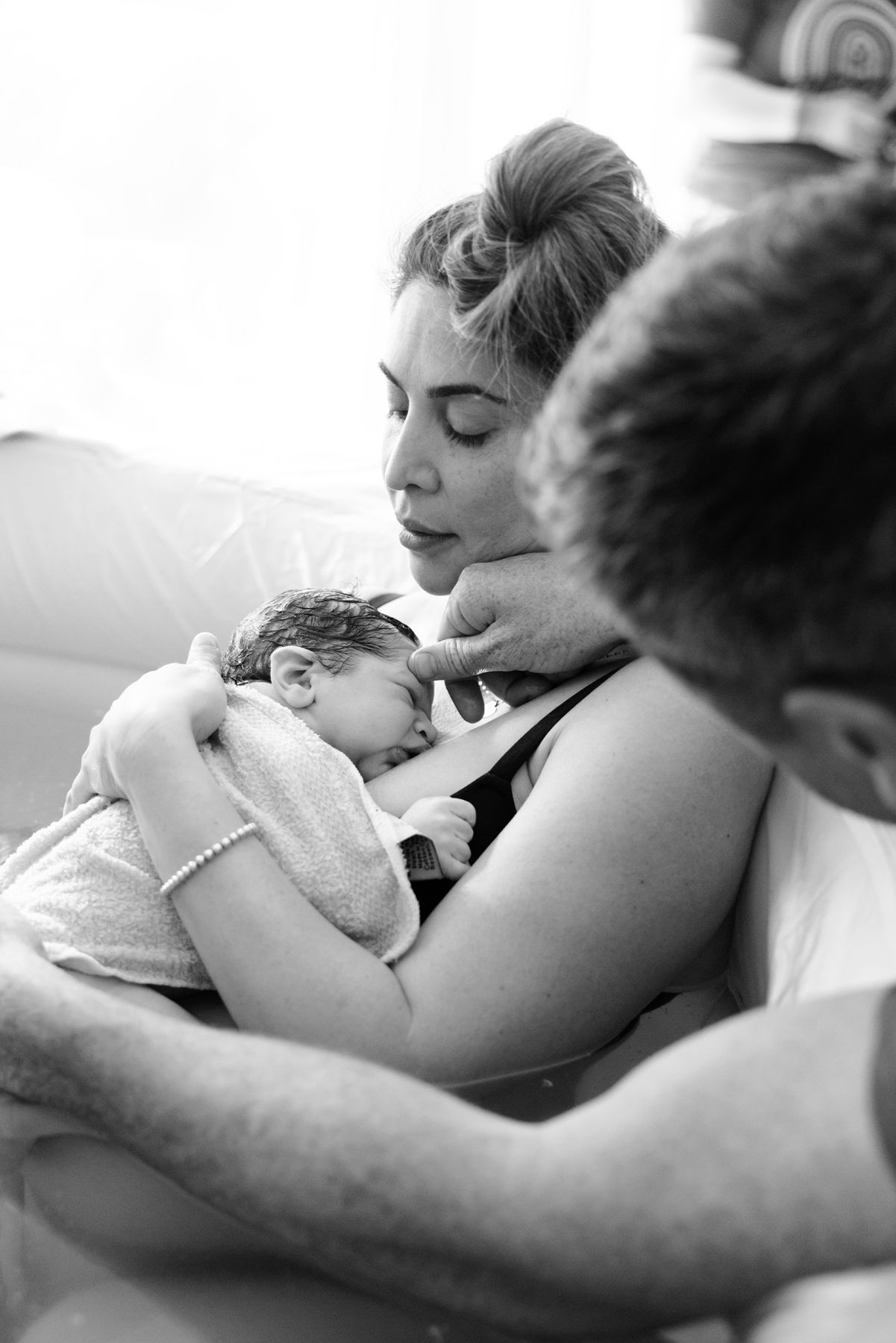 New parents share intimate moment gazing at sleeping newborn wrapped in white towel after birth.