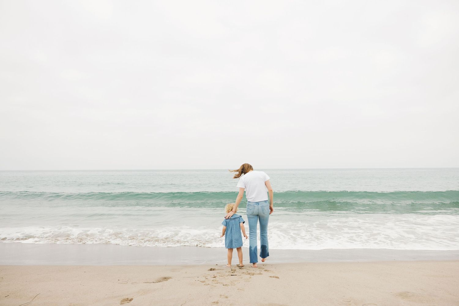 Adult and child walking hand-in-hand toward ocean waves on a sandy beach under an overcast sky.