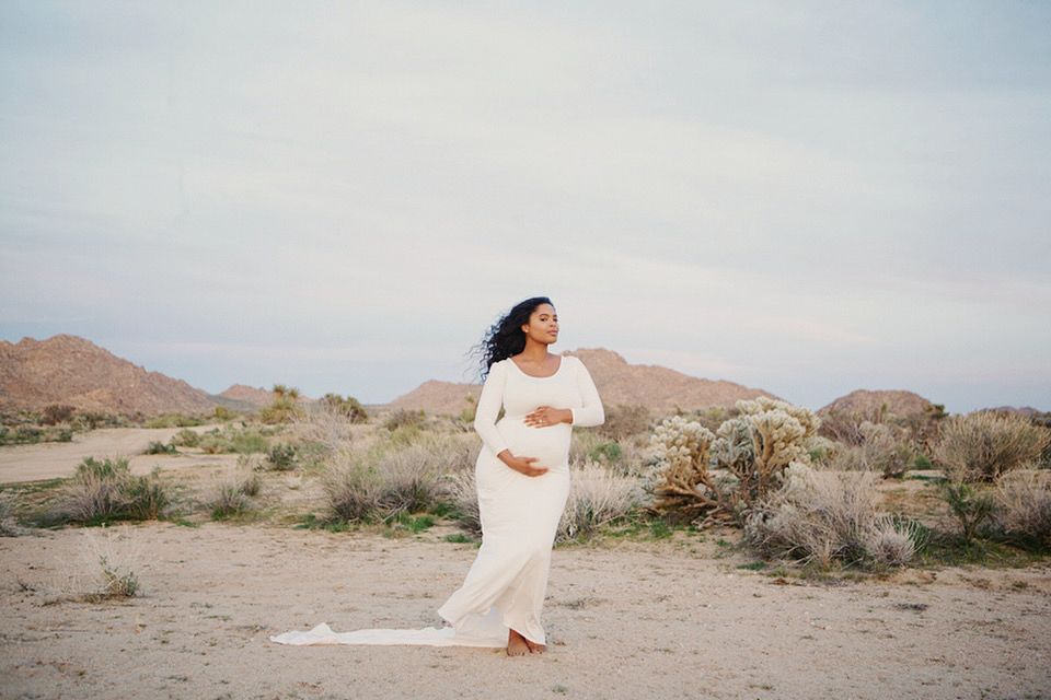 Pregnant woman in white gown poses in desert landscape with mountains and shrubs in background.