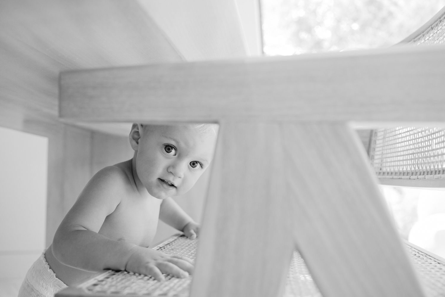 Black and white photo of a curious baby peeking through wooden furniture legs, looking up with wide eyes.