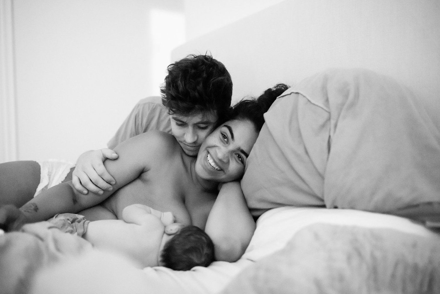 Black and white photo of couple smiling together in bed with newborn baby.