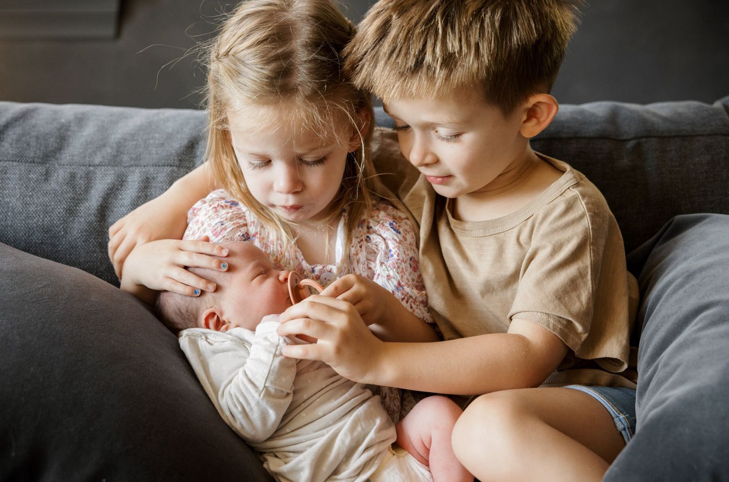 Two young siblings gently hold and admire their newborn baby on a gray couch.