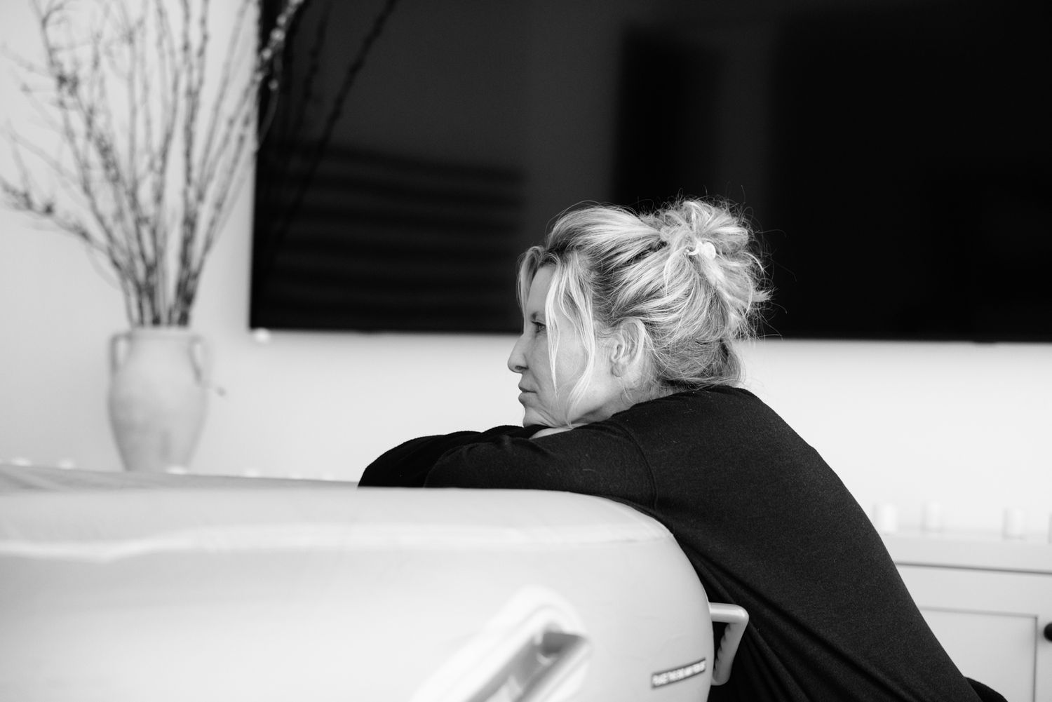 A blonde woman in black rests her arms on the edge of a birthing tub during a home water birth.