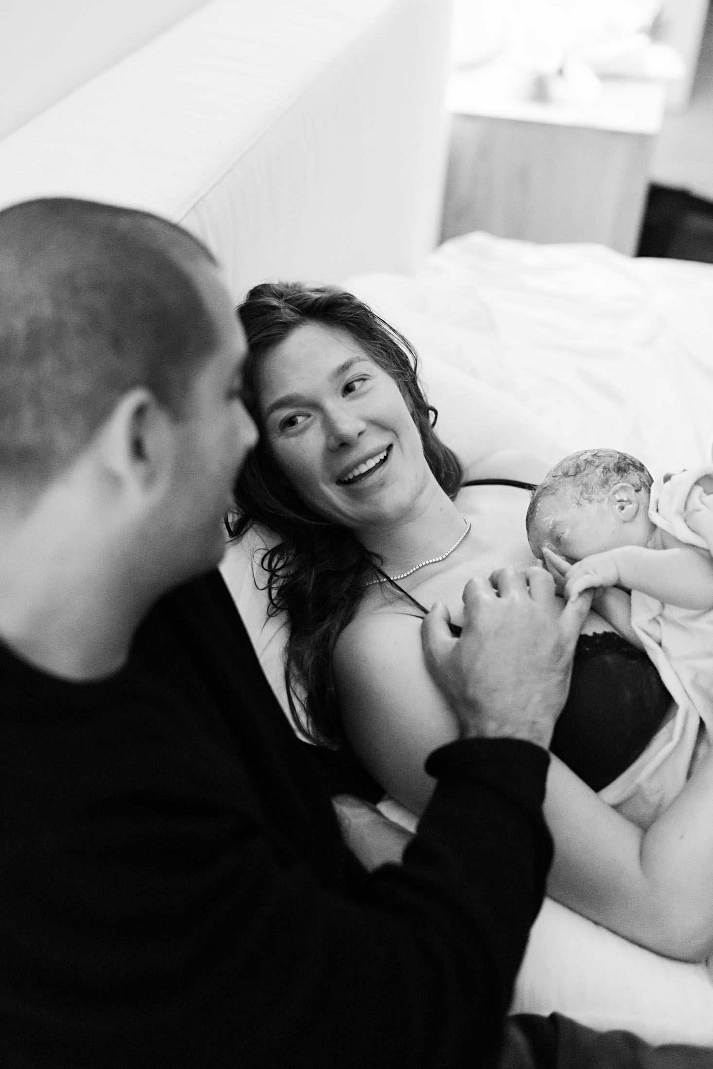 Smiling new mother holds newborn baby while father reaches out during home birth.