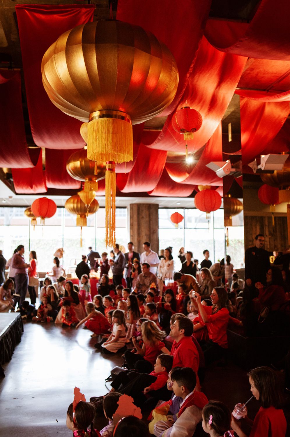 A large crowd sits beneath colorful red and gold Chinese lanterns at an indoor Lunar New Year celebration event.