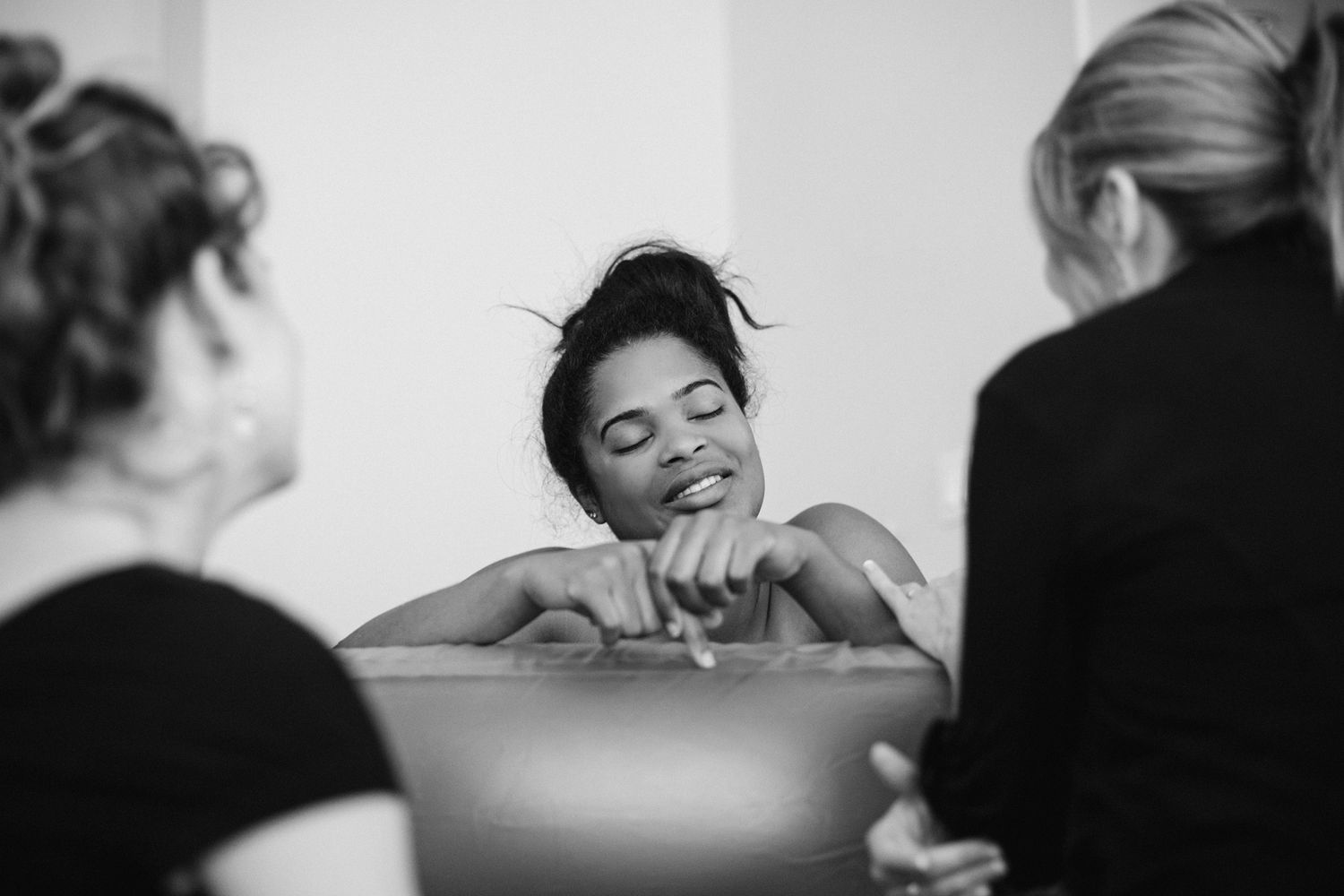 Black and white photo of a woman relaxing in a bath, attended by two caregivers in a serene setting.