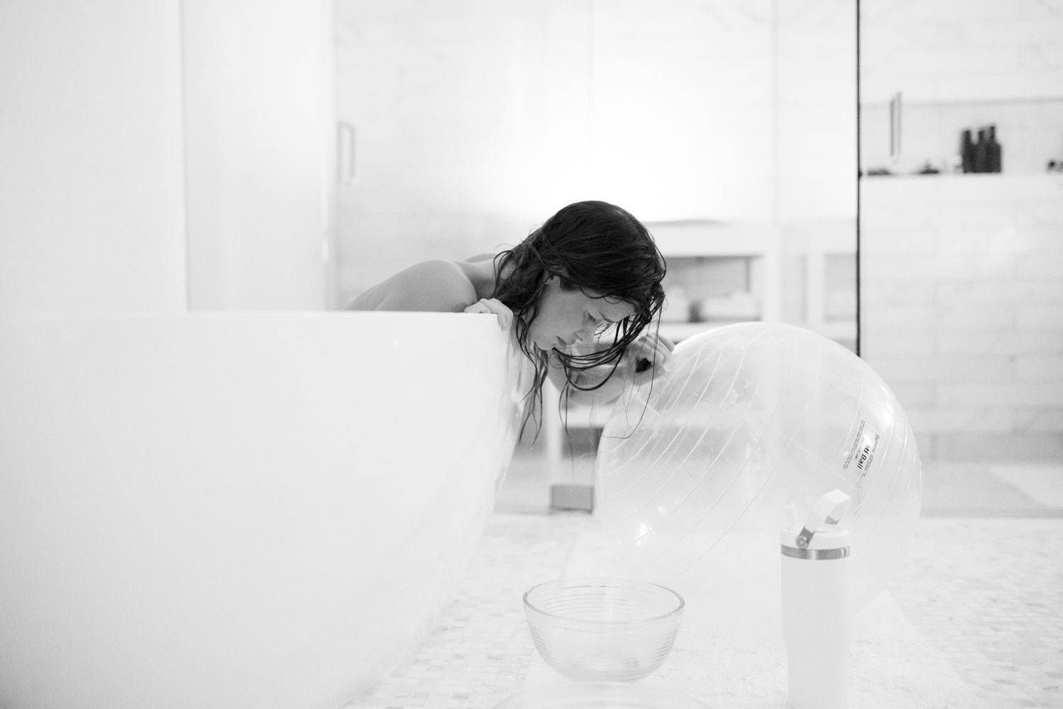 Black and white photo of a woman leaning over a bathtub in a bright white bathroom with a bowl nearby.