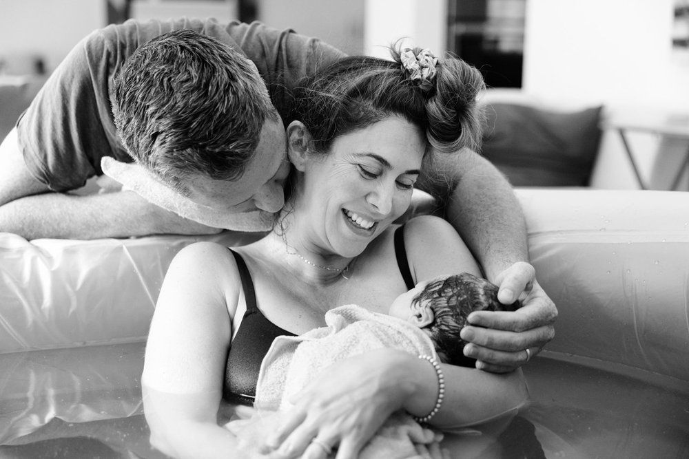 Parents joyfully holding newborn baby together in birth pool, black and white photo.