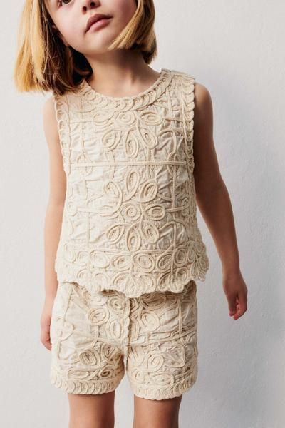 Young girl wearing a cream lace sleeveless top and matching shorts set with floral embroidery detail.