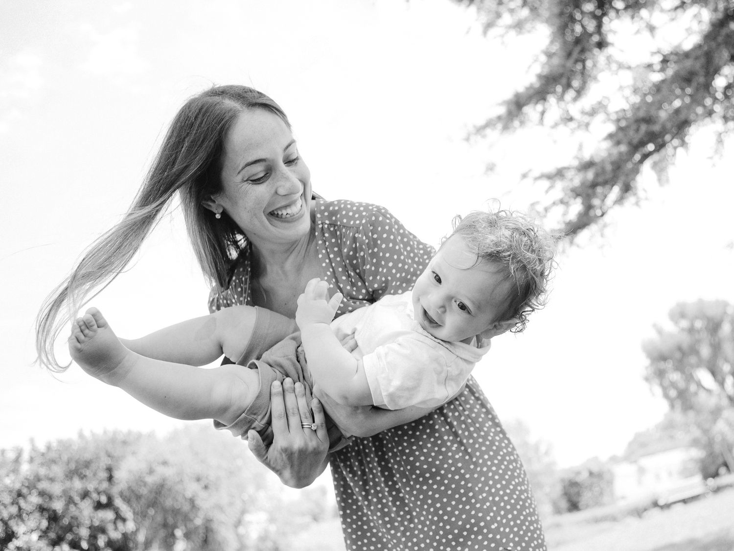 Black and white photo of a smiling mother joyfully holding her laughing baby outdoors in a sunny park.