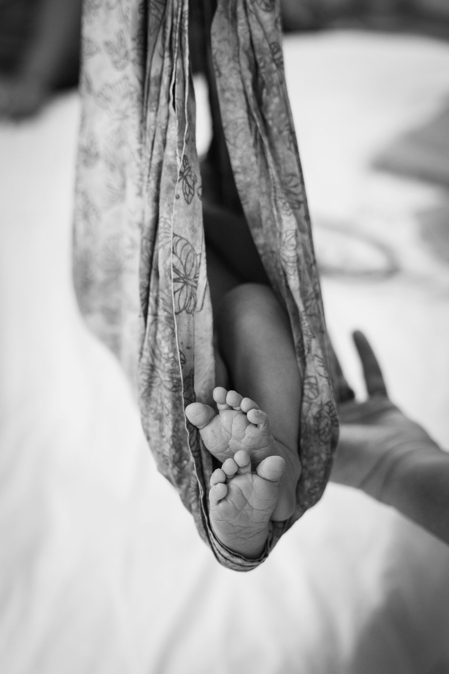 Black and white photo of a newborn baby resting in a floral fabric sling, tiny feet visible in the foreground.