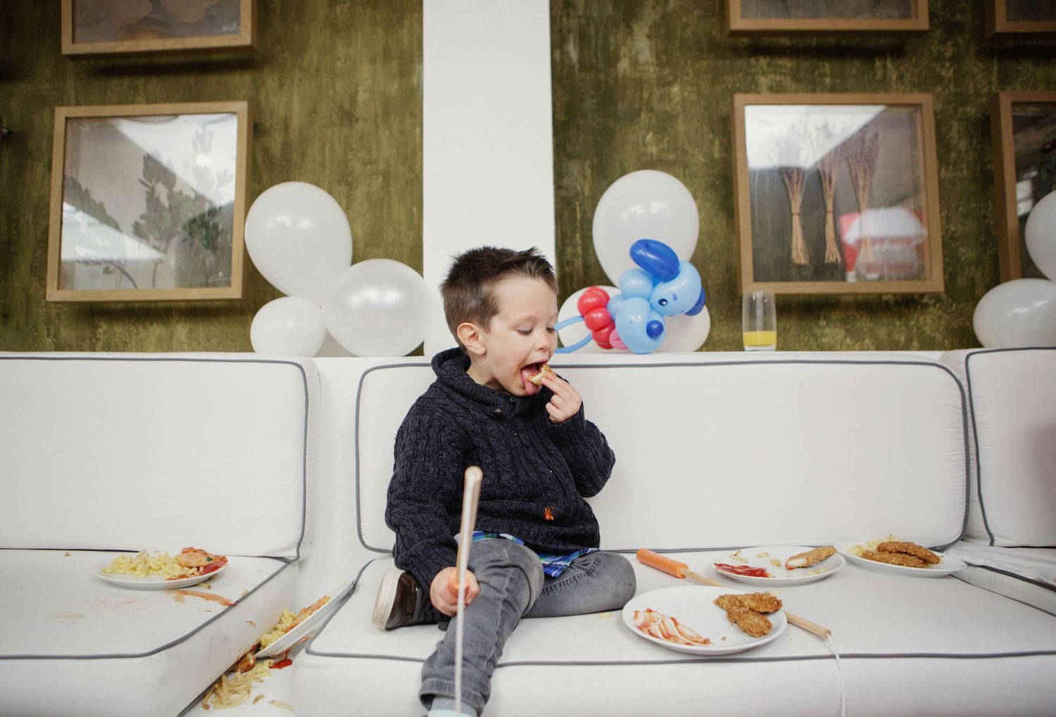 Young toddler boy eating cake at birthday party sitting on white sofa surrounded by balloons.