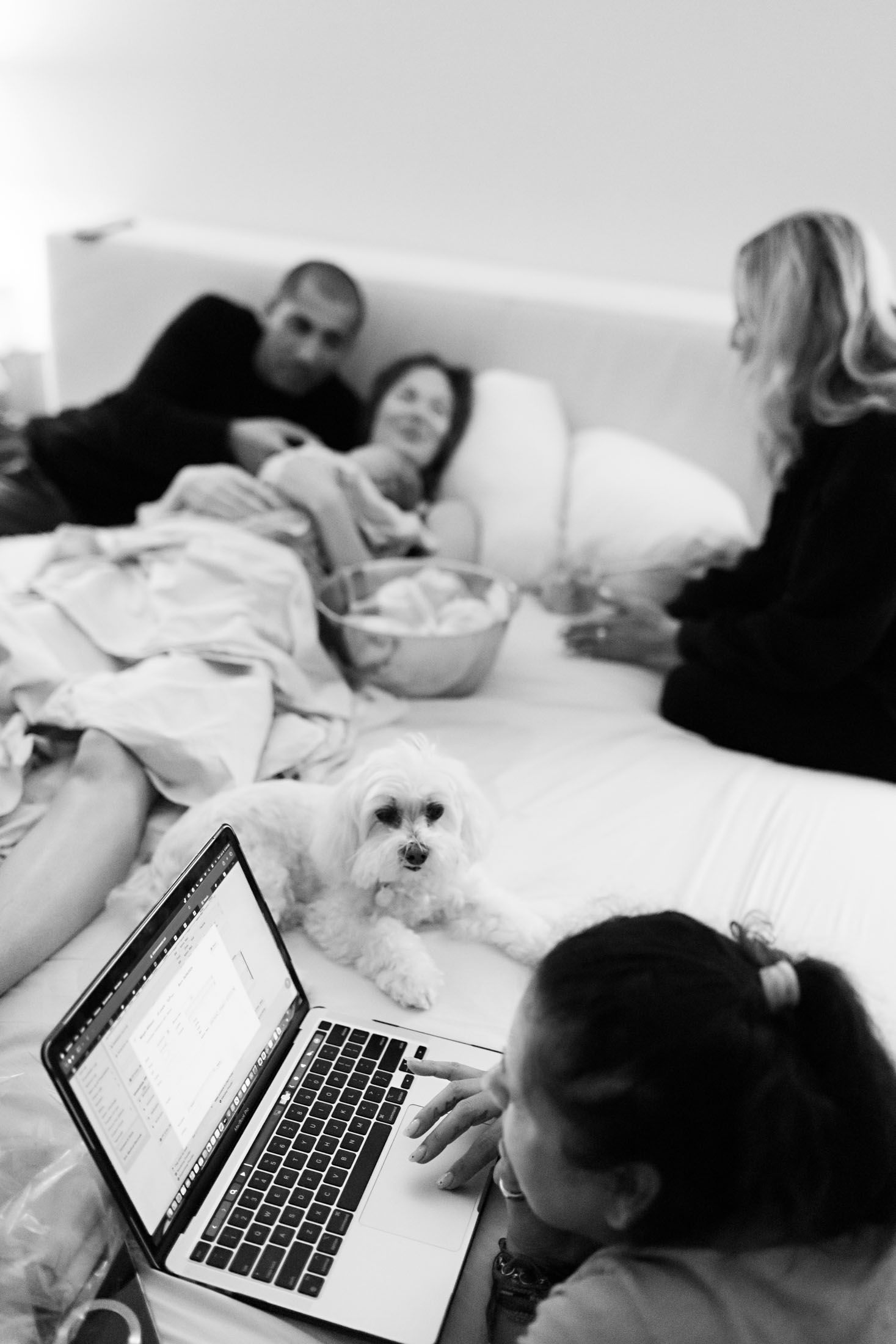 Family relaxes on bed with small white dog and laptop after home birth.